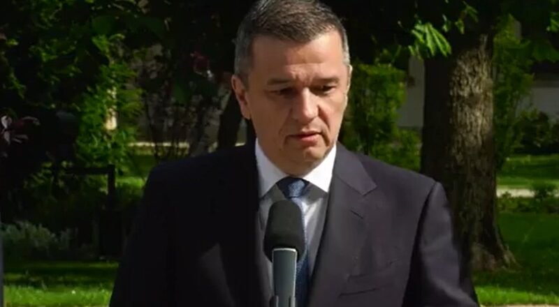 Man in a dark suit speaks into a microphone outdoors, with trees in the background.