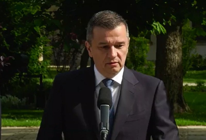 Man in a dark suit speaks into a microphone outdoors, with trees in the background.