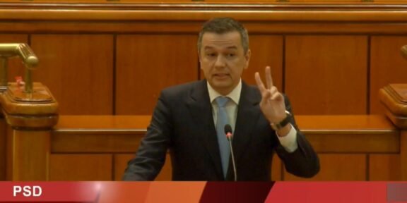 Male politician in a dark suit speaks at a wooden parliamentary podium, raising three fingers; PSD banner and name Grindeanu Sorin-Mihai visible on screen