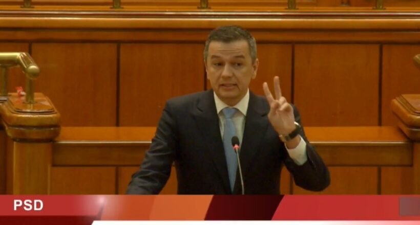 Male politician in a dark suit speaks at a wooden parliamentary podium, raising three fingers; PSD banner and name Grindeanu Sorin-Mihai visible on screen