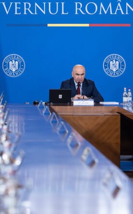 Bald man speaks at a central podium in a government meeting, with a long conference table and Romanian and EU flags in a blue backdrop behind him.