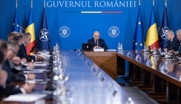 Bald man speaks at a central podium in a government meeting, with a long conference table and Romanian and EU flags in a blue backdrop behind him.