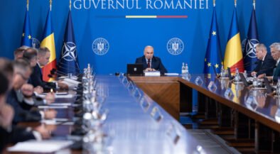 Bald man speaks at a central podium in a government meeting, with a long conference table and Romanian and EU flags in a blue backdrop behind him.