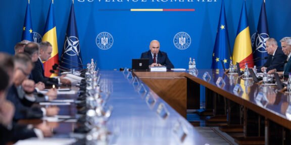 Bald man speaks at a central podium in a government meeting, with a long conference table and Romanian and EU flags in a blue backdrop behind him.