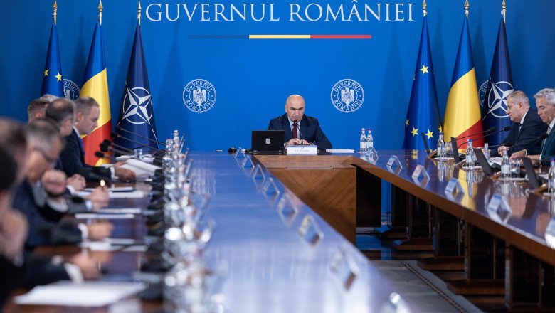 Bald man speaks at a central podium in a government meeting, with a long conference table and Romanian and EU flags in a blue backdrop behind him.