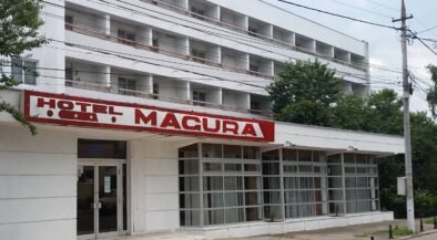 Exterior of a white multi-story hotel with a red 'HOTEL MACURA' sign above a glass-front entrance.