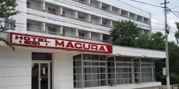 Exterior of a white multi-story hotel with a red 'HOTEL MACURA' sign above a glass-front entrance.