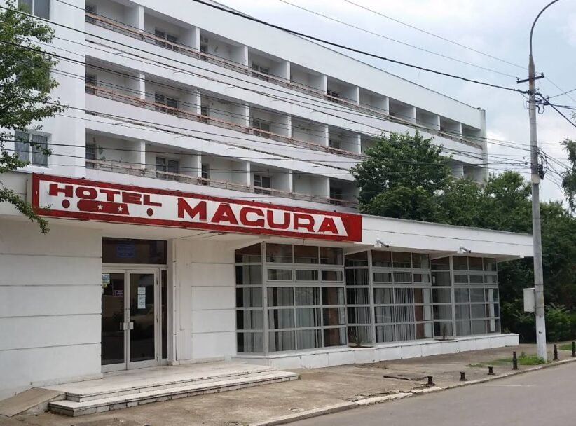 Exterior of a white multi-story hotel with a red 'HOTEL MACURA' sign above a glass-front entrance.