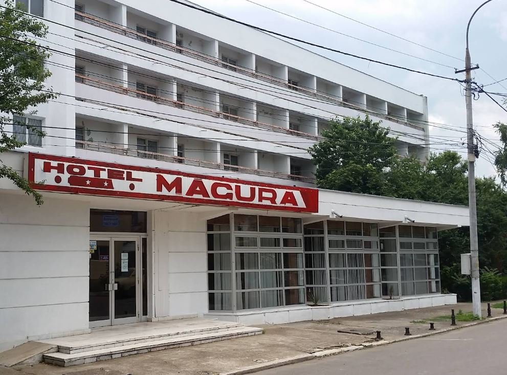 Exterior of a white multi-story hotel with a red 'HOTEL MACURA' sign above a glass-front entrance.