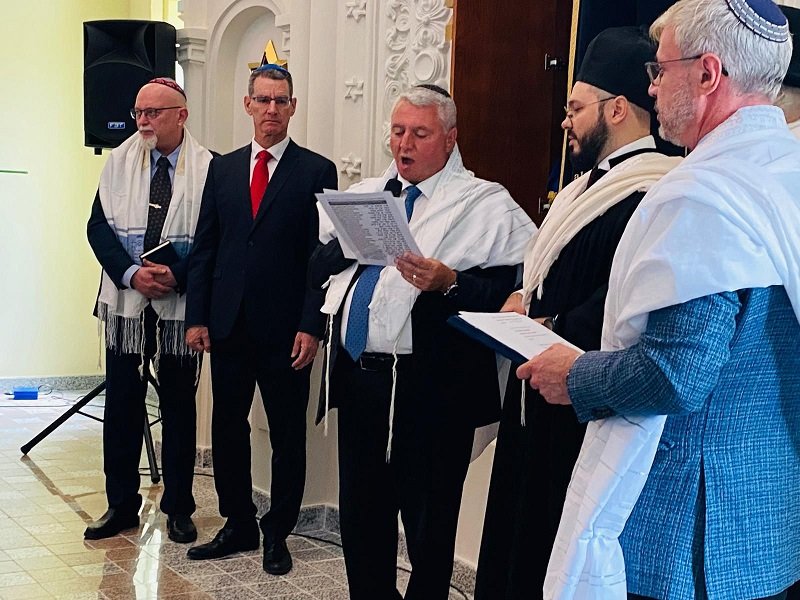Man in a suit reads from a sheet as four other men in white prayer shawls stand nearby in a formal ceremony setting.