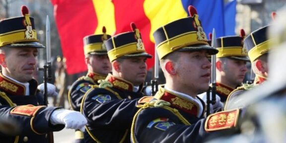 Group of uniformed soldiers marching with rifles during a parade, with red, yellow, and blue flags in the background.