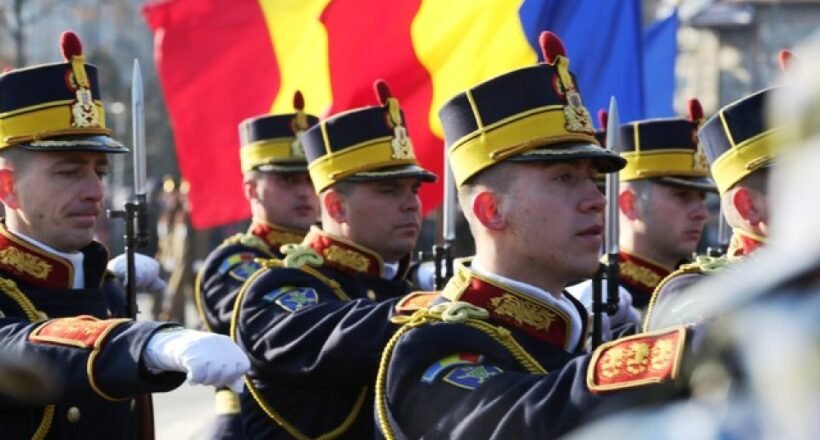 Group of uniformed soldiers marching with rifles during a parade, with red, yellow, and blue flags in the background.