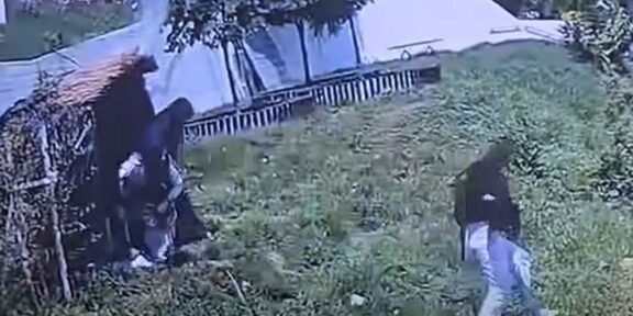 Two people in a grassy yard near a wooden fence and a white tent; one person stands by the fence while another walks uphill through tall grass