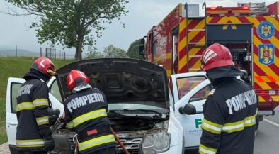 Three firefighters in turnout gear inspect the engine of a white van with its hood open on a roadside, a red fire truck in the background