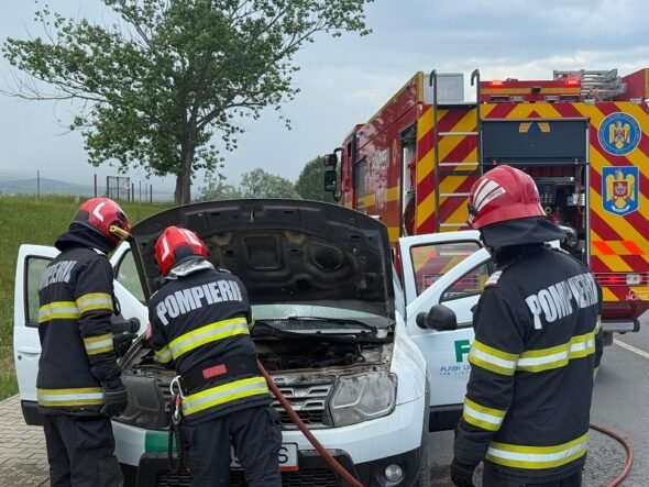 Three firefighters in turnout gear inspect the engine of a white van with its hood open on a roadside, a red fire truck in the background