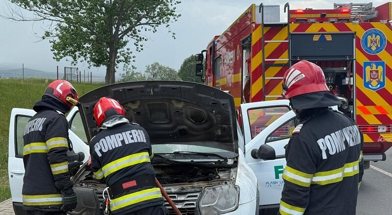 Three firefighters in turnout gear inspect the engine of a white van with its hood open on a roadside, a red fire truck in the background