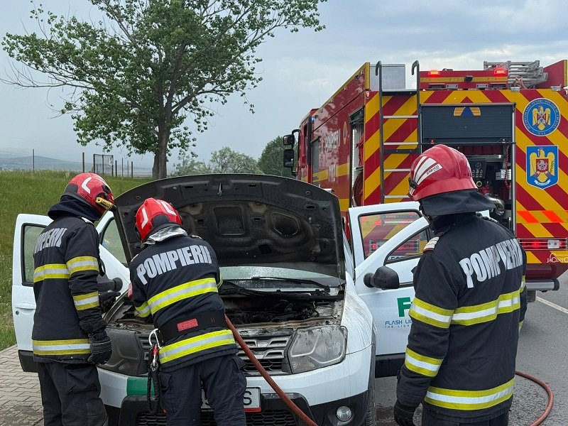 Three firefighters in turnout gear inspect the engine of a white van with its hood open on a roadside, a red fire truck in the background