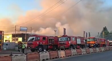 Several red fire trucks parked along a street as thick smoke and flames rise from a building; firefighters are on scene.