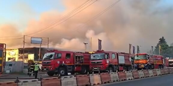 Several red fire trucks parked along a street as thick smoke and flames rise from a building; firefighters are on scene.