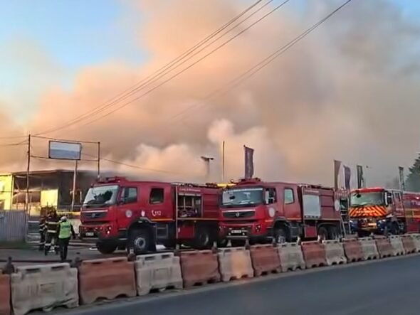 Several red fire trucks parked along a street as thick smoke and flames rise from a building; firefighters are on scene.