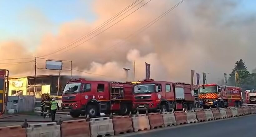 Several red fire trucks parked along a street as thick smoke and flames rise from a building; firefighters are on scene.