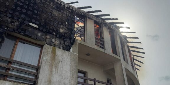 Two-story house with a severely burnt roof and exposed beams after a fire, soot on the walls you can see through the windows.
