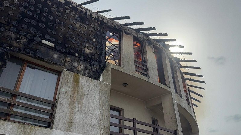 Two-story house with a severely burnt roof and exposed beams after a fire, soot on the walls you can see through the windows.