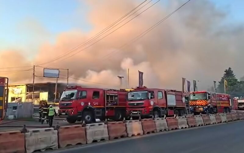 Several red fire trucks parked along a street as thick smoke and flames rise from a building; firefighters are on scene.