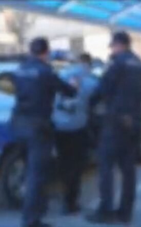 Two police officers restraining a person beside a blue police SUV under a covered walkway outside a building.