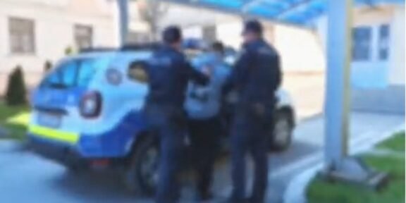 Two police officers restraining a person beside a blue police SUV under a covered walkway outside a building.
