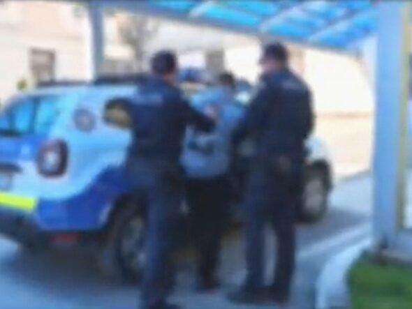 Two police officers restraining a person beside a blue police SUV under a covered walkway outside a building.