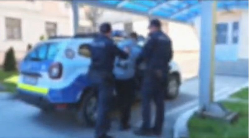 Two police officers restraining a person beside a blue police SUV under a covered walkway outside a building.