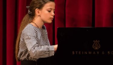 Girl with long hair in a checkered blouse playing a Steinway piano on a stage with red curtains, focused on the keys.