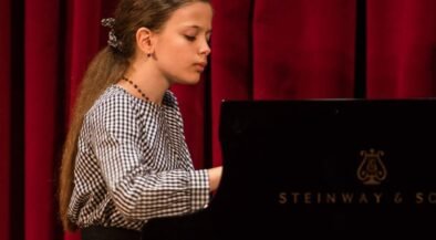 Girl with long hair in a checkered blouse playing a Steinway piano on a stage with red curtains, focused on the keys.