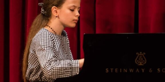 Girl with long hair in a checkered blouse playing a Steinway piano on a stage with red curtains, focused on the keys.