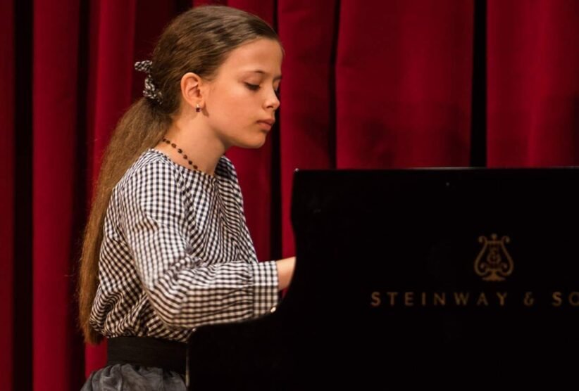 Girl with long hair in a checkered blouse playing a Steinway piano on a stage with red curtains, focused on the keys.