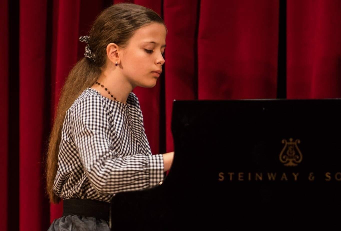 Girl with long hair in a checkered blouse playing a Steinway piano on a stage with red curtains, focused on the keys.
