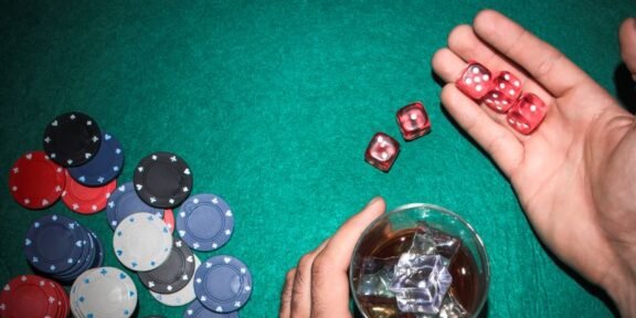 Casino table scene with colorful poker chips piled on the left, red dice in a player's hand, and a glass with ice on the right.