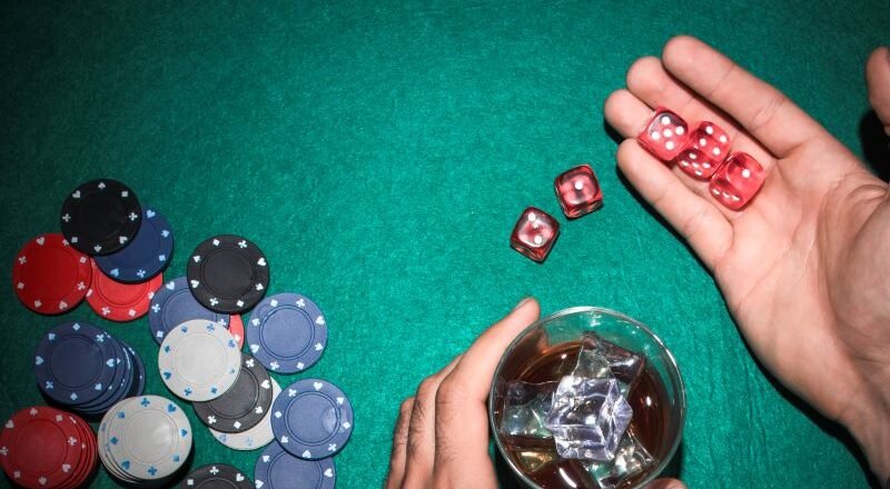 Casino table scene with colorful poker chips piled on the left, red dice in a player's hand, and a glass with ice on the right.