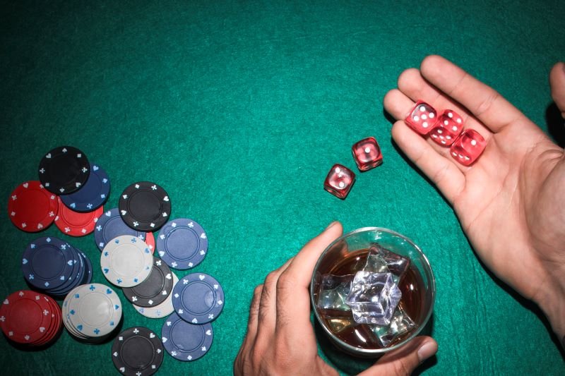 Casino table scene with colorful poker chips piled on the left, red dice in a player's hand, and a glass with ice on the right.