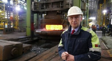 Industrial foreman in hard hat and safety jacket beside a glowing molten metal furnace in a factory.