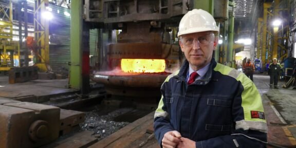 Industrial foreman in hard hat and safety jacket beside a glowing molten metal furnace in a factory.