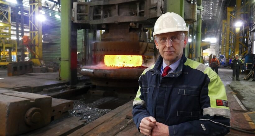 Industrial foreman in hard hat and safety jacket beside a glowing molten metal furnace in a factory.
