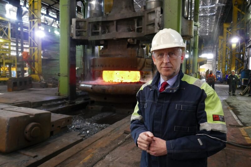 Industrial foreman in hard hat and safety jacket beside a glowing molten metal furnace in a factory.