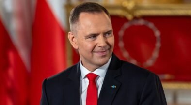 Smiling man in a dark suit and red tie in a formal room with red and gold decor in the background.