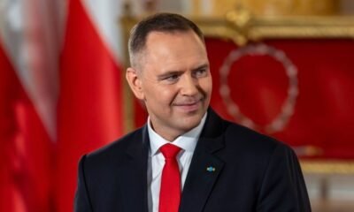 Smiling man in a dark suit and red tie in a formal room with red and gold decor in the background.