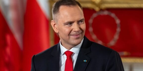 Smiling man in a dark suit and red tie in a formal room with red and gold decor in the background.