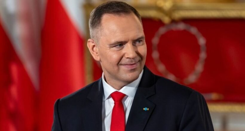 Smiling man in a dark suit and red tie in a formal room with red and gold decor in the background.
