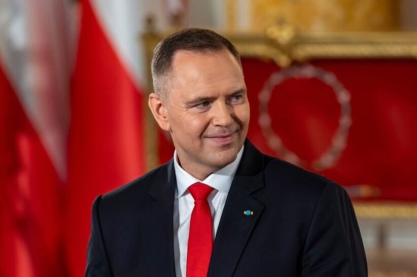 Smiling man in a dark suit and red tie in a formal room with red and gold decor in the background.