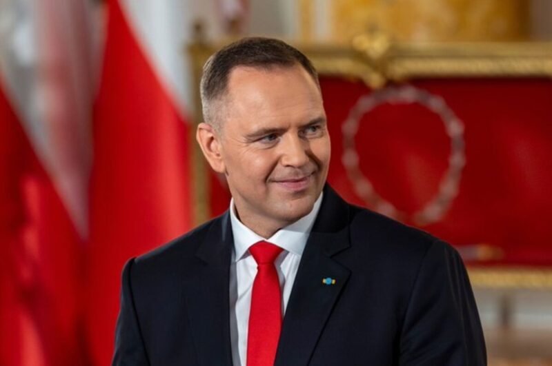 Smiling man in a dark suit and red tie in a formal room with red and gold decor in the background.